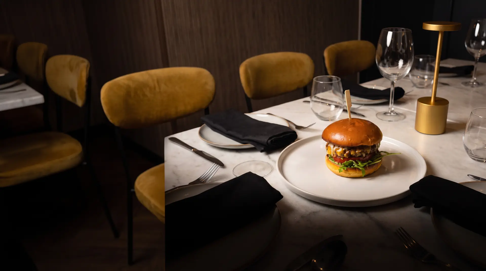 Mil Estrellas Burger en plato blanco sobre mesa de mármol con copas de vino y sillas de terciopelo mostaza