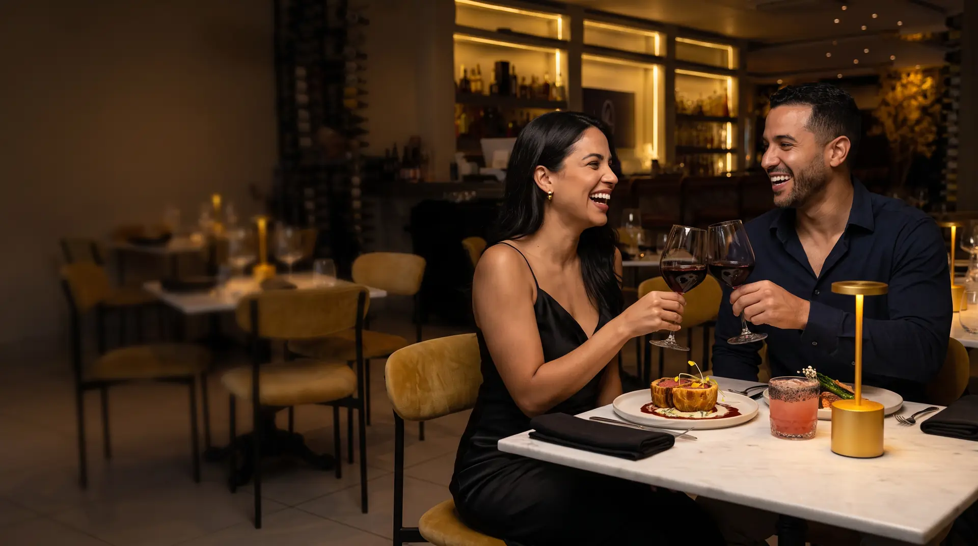 Pareja brindando con vino tinto en una mesa de mármol de Mil Estrellas, con beef Wellington y un cóctel Smokiss, y la barra iluminada al fondo